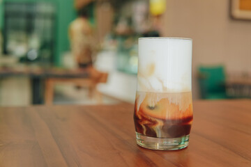 A glass of iced coffee with creamy milk foam latte on top part, blurred cafe interior background.