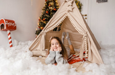 A charming girl, nestled in a cozy Christmas teepee, smiles happily. The soft white snow and...