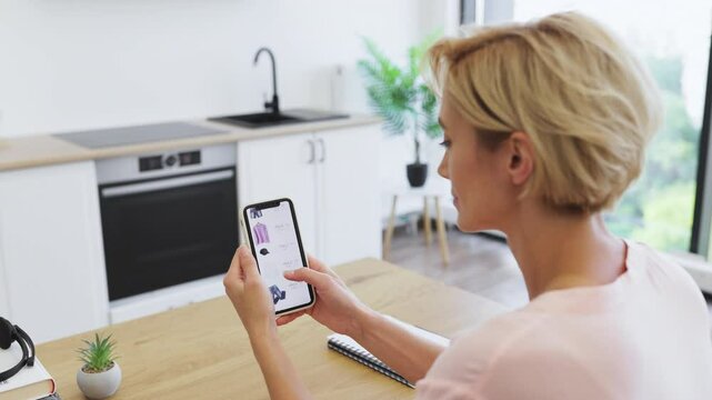 Caucasian woman in her 30s sitting at kitchen table using smartphone for online shopping. Casual attire and bright, airy kitchen setting convey relaxing and modern lifestyle.