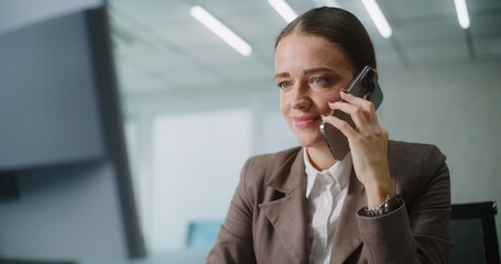 Positive female marketing specialist or E-commerce manager talks to business partner, consults client on mobile phone, uses computer. Caucasian businesswoman works in bright modern office. E-business.