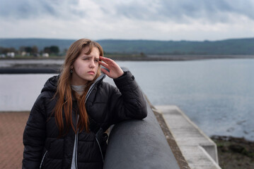 A young teen girl in jacket gazes thoughtfully over tranquil seaside view. Her expression suggests...