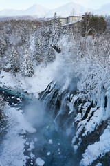 Majestic Winter Waterfall Surrounded by Snowy Pines and Ice-Covered Rocks with Mist Rising in a Serene Landscape at Dawn