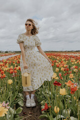 A girl in a yellow dress and apron with a water can in a red and green tulip flower field on a gloomy rainy cloudy sky background happily running around. 