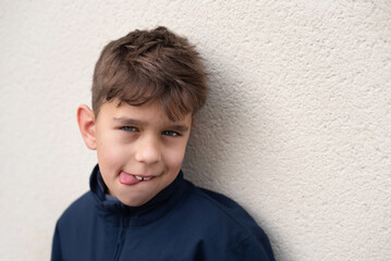 A young boy in a navy jacket playfully sticks out his tongue against a textured cream wall,...
