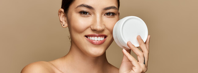 Young woman smiles radiantly while demonstrating skincare product in calm, neutral backdrop.