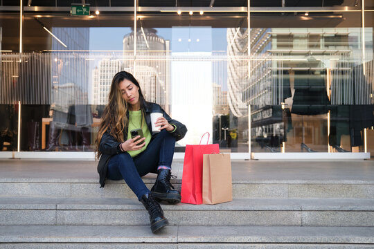 Young woman using phone and drinking coffee after shopping in the city