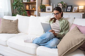 Young design professional business woman sitting on sofa at home using digital tablet. Female choosing new wardrobe from internet store for online shopping and ordering.