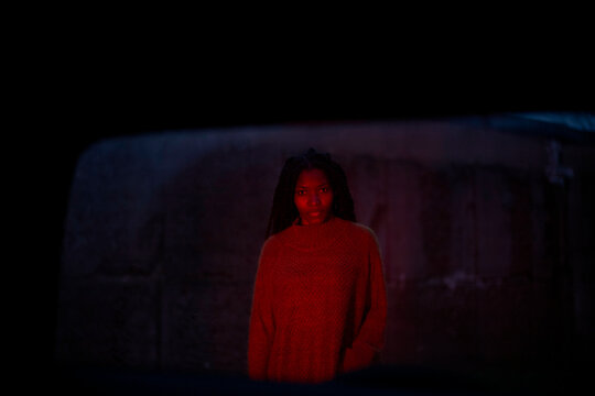 Woman in red sweater standing in the dark behind a car, illuminated by a soft red light, looking at the camera, Brandenburg, Germany
