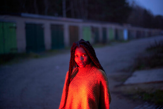Woman with braided hair in a red sweater standing outdoors at twilight with a row of green doors in the background, Brandenburg, Germany