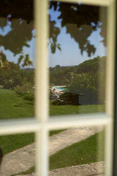 View of a person lounging by a pool reflected in a window pane framed by lush greenery, Portugal