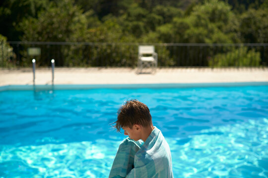 Young boy wrapped in a towel standing by a clear blue swimming pool, with a backdrop of green foliage, Portugal