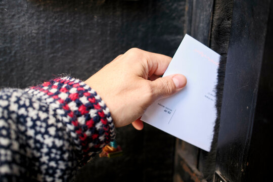 A close-up view of a hand holding a white envelope as it is being inserted into a dark mail slot.