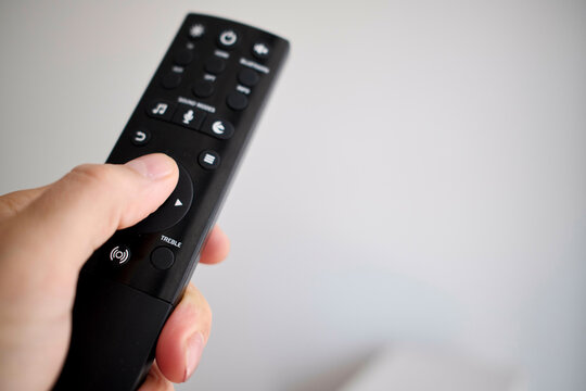 Close-up of a hand holding a black television remote control against a blurred white background.