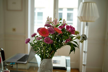 Fresh colorful flowers in a white vase on a table with a laptop and lamp in a bright room.