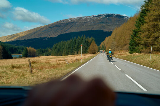 A scenic drive with views of a motorcyclist on a road bordered by pine trees and mountains under a clear blue sky, Highlands, Scotland