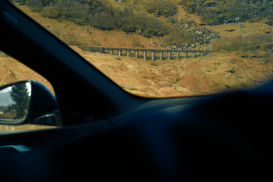 View of an ancient stone bridge through a car window framed by the interior of the vehicle against a backdrop of rugged terrain, Highlands, Scotland