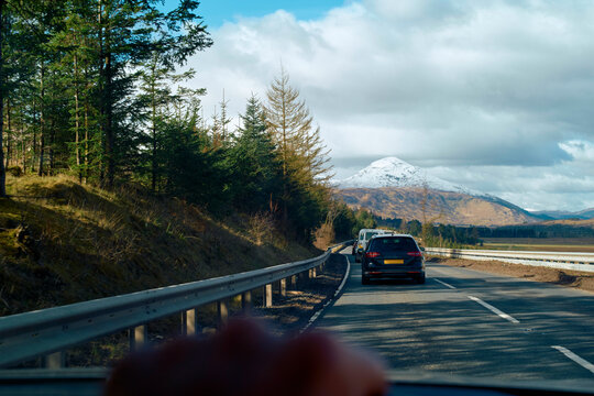 Car driving on a road with a snow-capped mountain in the background and pine trees lining the route under a blue sky with clouds, Highlands, Scotland - Powered by Adobe