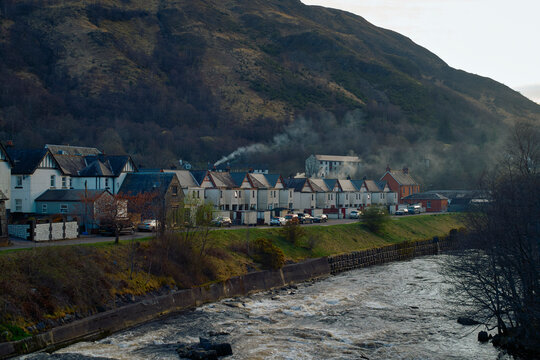Early morning view of a 0uaint European village by a flowing river with smoke rising from a chimney against a backdrop of hills, Scotland