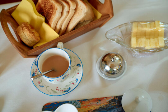 A cozy breakfast setup featuring a cup of tea with milk, sliced bread in a basket, butter, and a lit candle on a white tablecloth, Scotland