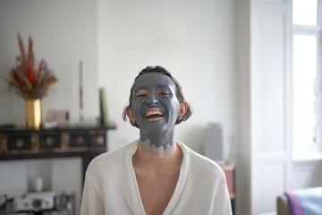 Young woman with a facial mask smiling in a bright, cozy room.