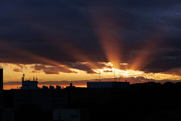 Brilliant orange sun rays stretch across dark blue clouds partially blanketing the sunset sky....