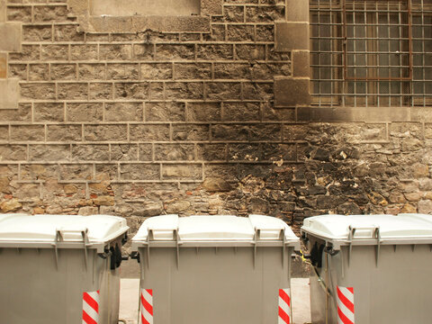 Three gray garbage bins lined up against an old stone wall with a metal grid and burning traces above them, Barcelona, Spain