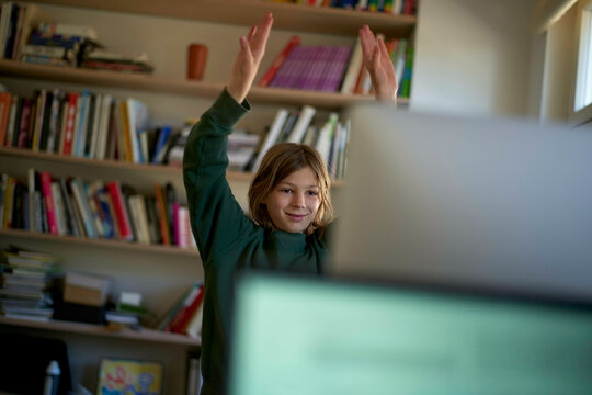 Smiling child with arms raised in a victory gesture in front of a computer at a desk with a bookshelf in the background.