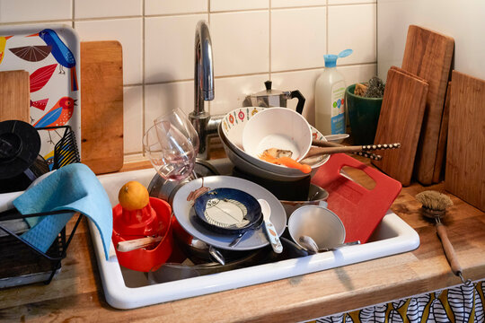 A cluttered kitchen sink filled with dirty dishes, including pots, pans, utensils, and glassware, Germany
