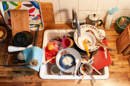 A sink full of dirty dishes and kitchen utensils with colorful designs, surrounded by a wood countertop and backsplash, Germany