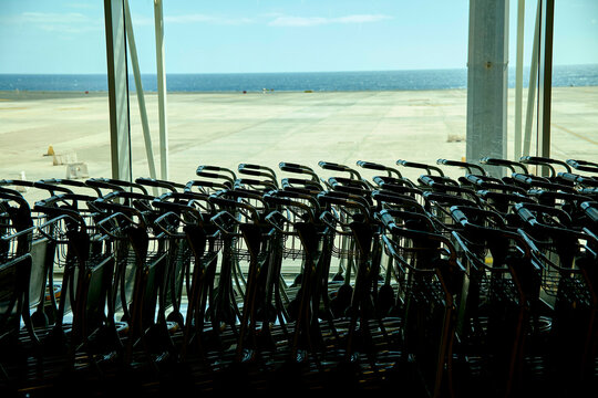 Rows of empty black trolleys facing a large window overlooking an airport runway under a clear blue sky, Fuerteventura, Spain