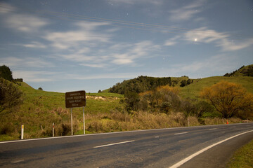 Nighttime landscape of a winding road with a sign indicating a nearby chain reserve and sheep grazing under a starry sky, Northland, New Zealand
