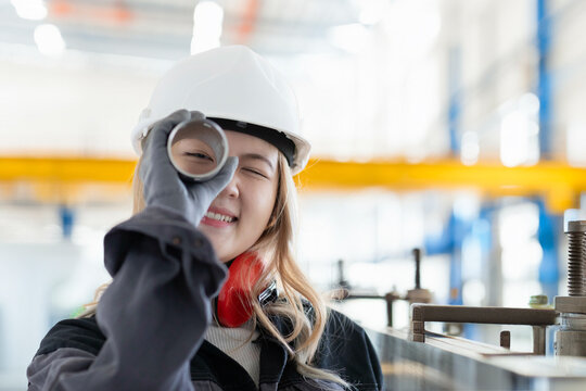 Smiling female engineer with hard hat looking through a metal pole in an industrial setting, Germany