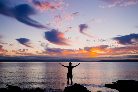 Silhouetted woman standing with arms raised against a vibrant sunrise over the ocean. - Powered by Adobe