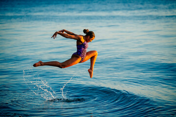 Woman in purple swimwear joyfully leaps above the water at the beach during sunset, USA