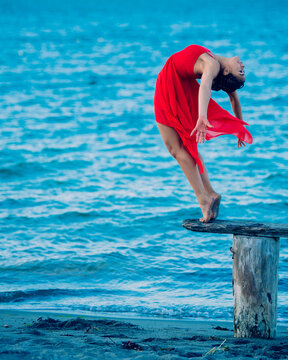 Woman in a red dress performing an elegant dance move on a wooden stump by the sea, USA