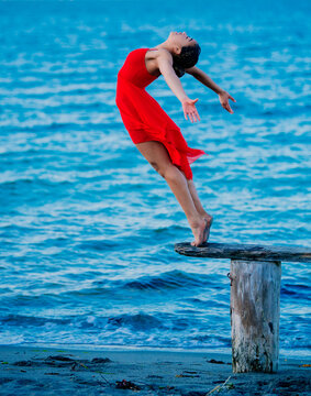 Graceful dancer in a red dress performing a dance move on a wooden stump by the blue sea, USA