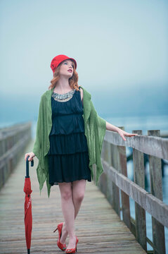 Elegant woman in a red beret and heels holding an umbrella while standing on a wooden pier shrouded in fog.