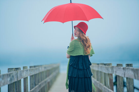 A woman with red hair holding a red umbrella stands on a foggy pier looking over her shoulder.