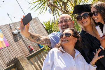 A joyful group takes a selfie to celebrate a graduation, with two smiling women in graduation caps and a tattooed man wearing glasses, Bournemouth, Dorset UK