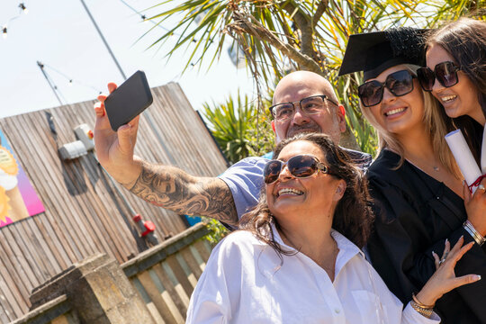 A joyful group of four taking a selfie on a sunny day, with two individuals in graduation caps celebrating a milestone, Bournemouth, Dorset UK