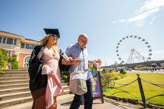 Female graduate in cap and gown walking with dad in casual attire, with a large Ferris wheel in the background, Bournemouth, Dorset UK