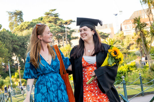 Two smiling women walking in a park, one in a graduation cap and gown holding a sunflower bou0uet, Bournemouth, Dorset UK