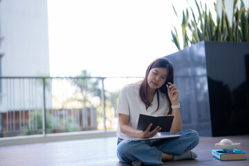 Fototapeta premium Young asian woman sitting cross-legged on a balcony floor, reading from a notebook while adjusting her glasses, enjoying a moment of relaxation and tranquility in her cozy apartment