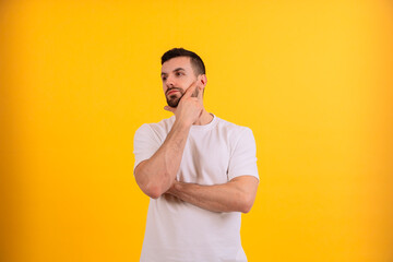 A young man is standing against a bright yellow background, looking up thoughtfully with a hand on...