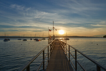 Obraz premium A serene sunset over a calm river, with vibrant orange and pink hues in the sky. A small sailboat is anchored in the water, reflecting the colorful sky.