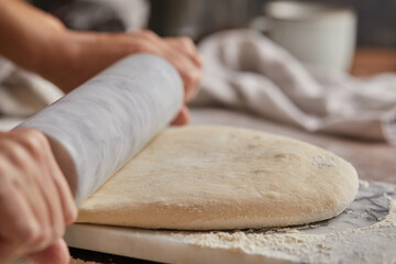 Cooking homemade bread. Sieving flour over raw bread dough rolled with rolling pin on marble board, dark background, selective focus. Baker hands on dough.