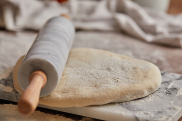 Cooking homemade bread. Sieving flour over raw bread dough rolled with rolling pin on marble board, dark background, selective focus. Baker hands on dough.