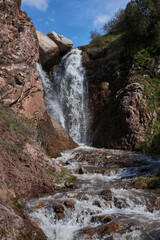 Small waterfall between stones and rocky slopes, blue sky. Summer natural landscape, Kyrgyzstan