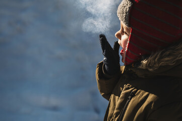 Fototapeta premium 4 year old boy in winter hat and jacket inhales frosty air and exhales steam. breathe out steam outdoor in winter