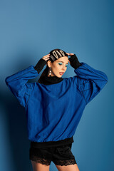 A fashionable young woman poses confidently in stylish attire against a blue backdrop.